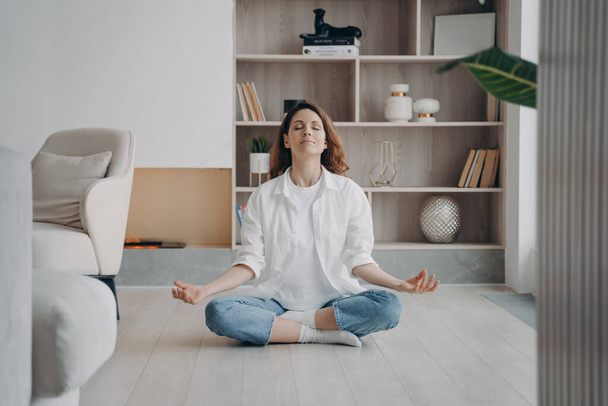 Donna calma seduta in posizione loto con gesto mudra sul pavimento in soggiorno moderno. Le donne pacifiche meditano, praticano yoga, respirano aria fresca da sole a casa. Ricreazione, stile di vita sano. - Foto, immagini