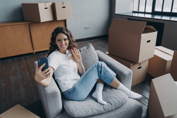 スマートフォンのグリーティングフォロワーが手を振っている幸せな女性の住宅所有者ブロガーは、移転日に所持品と段ボール箱の近くに新しい家に座っている。移動と修理の概念. - 写真・画像