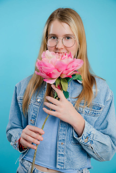 Csinos tini lány kezében nagy illatos rózsaszín friss virág hosszú szár üres kék háttér. Gyönyörű, szemüveges fiatal hölgy élvezi a virágzó növény illatát. Fényűző virágok koncepciója. - Fotó, kép
