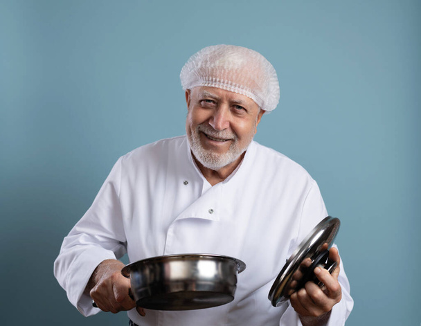 Vanhempi karismaattinen mieskokki uniformisissa astioissa ruoanlaittoon, kameran katseluun ja hymyilemiseen vaaleansinisellä taustalla - Valokuva, kuva