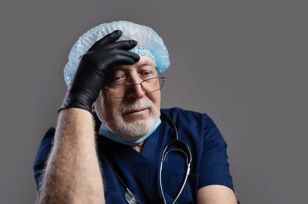 Un anciano doctor de pelo gris con un estetoscopio y una gorra y guantes negros, sostiene su cabeza, mira pensativamente hacia abajo, de cerca, fondo gris, espacio para el texto - Foto, imagen