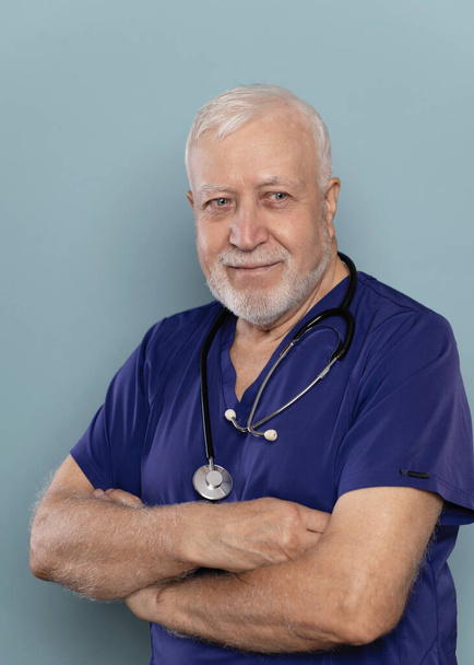 Un anziano medico dai capelli grigi con uno stetoscopio sta con le braccia incrociate sul petto su uno sfondo chiaro, guarda di lato, ritratto, primo piano - Foto, immagini