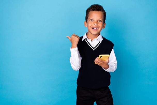 Photo of excited positive little guy dressed black vest pointing thumb empty space chatting modern device isolated blue color background. - Foto, Imagem