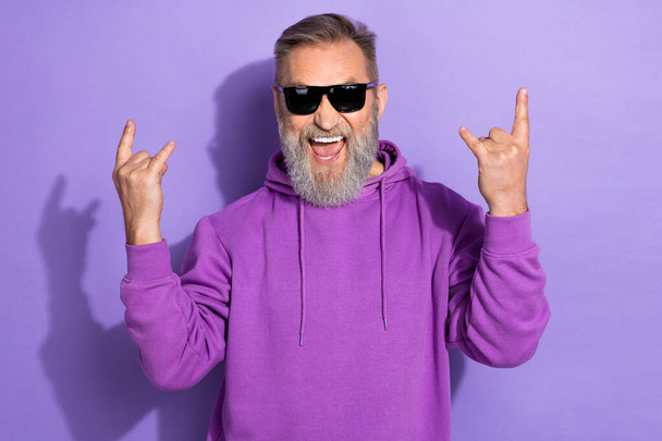 Фото красивого крутого пенсионера человека седой бороды, одетый в фиолетовую толстовку, показывает тяжелый металлический знак, выделенный на фиолетовом фоне. - Фото, изображение