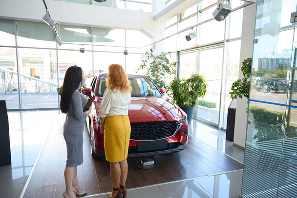 Gerente en vestido a cuadros y cliente en falda amarilla de pie delante de un modelo seleccionado de un coche rojo - Foto, Imagen