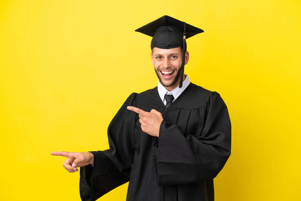 Молодой выпускник университета кавказский человек изолирован на желтом фоне удивлен и указал сторону - Фото, изображение