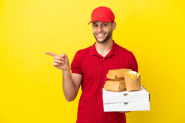 Pizzalieferant nimmt Pizzakartons und Burger über isoliertem Hintergrund entgegen und zeigt mit dem Finger zur Seite - Foto, Bild