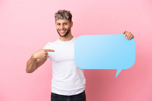 Young handsome caucasian man isolated on pink background holding an empty speech bubble and pointing it - Fotografie, Obrázek
