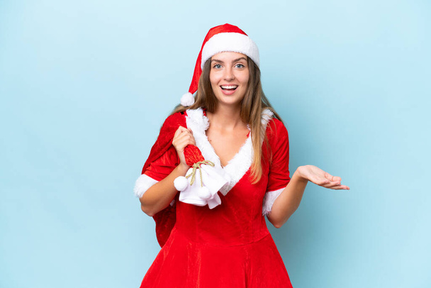 Anne Noel gibi giyinmiş genç beyaz kadın elinde Noel çuvalı tutarken mavi arka planda şok olmuş yüz ifadesi ile izole edilmiş. - Fotoğraf, Görsel