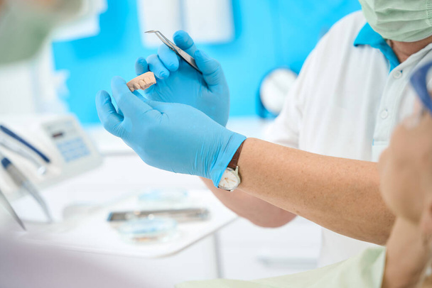 Retrato de vista recortada de las manos del médico en guantes azules con instrumento estéril y modelo de dientes en el centro médico - Foto, Imagen