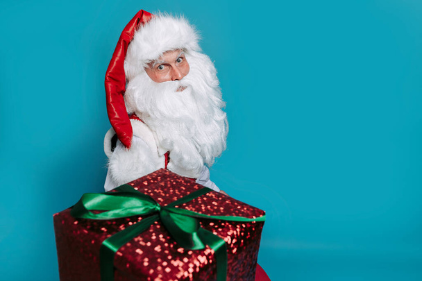 Photo of Santa Claus gives Christmas gift box while looking at camera on blue studio background. New Year concept  - Foto, Imagen
