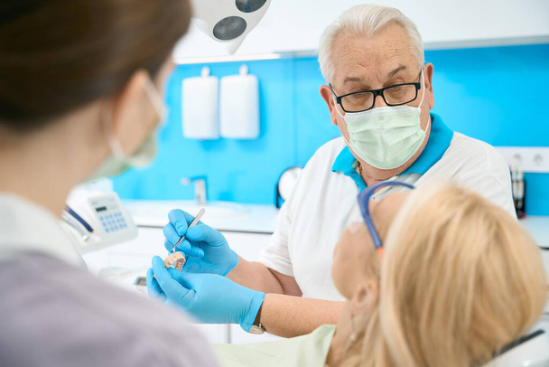 Крупный план портрет красивый мужчина врач в очках держит зубы модели и металлический инструмент во время обсуждения с пациентом в стоматологической клинике - Фото, изображение