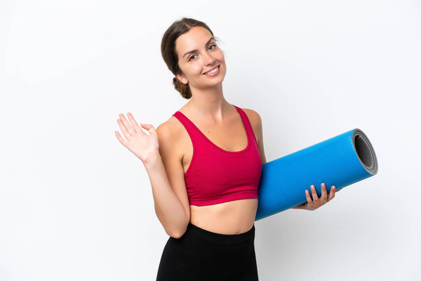 Young sport caucasian woman going to yoga classes while holding a mat isolated on white background saluting with hand with happy expression - Fotó, kép
