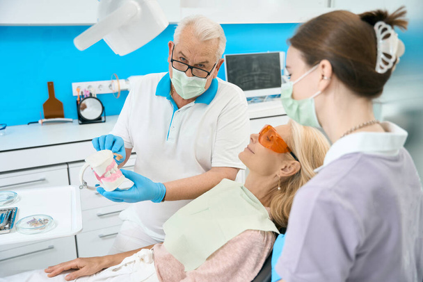 Taille op vooraanzicht portret van serieus tandarts wit kunstmatige kaak is het demonstreren van de juiste manier om tanden en tandvlees schoon te maken in tandheelkundige kliniek - Foto, afbeelding