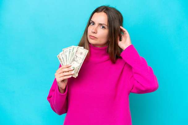 Nuori valkoihoinen nainen ottaa paljon rahaa eristetty sinisellä taustalla, joilla on epäilyksiä - Valokuva, kuva