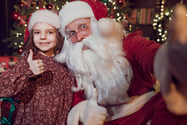 Santa Claus makes selfie with charming little girl who showing thumb up. Christmas background. - Fotoğraf, Görsel