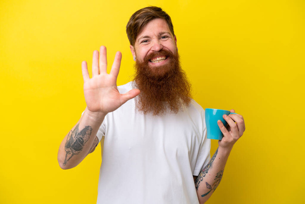 Rudowłosy mężczyzna z brodą trzymający kubek odizolowany na żółtym tle salutujący ręką ze szczęśliwym wyrazem twarzy - Zdjęcie, obraz