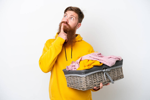 Rothaariger Mann mit Bart, der einen Wäschekorb auf weißem Hintergrund hält und beim Nachschauen Zweifel hat - Foto, Bild