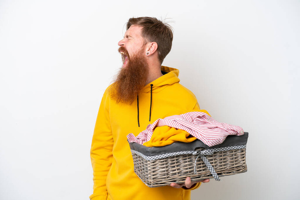 Rotschopf mit Bart hält einen Wäschekorb auf weißem Hintergrund und lacht in Seitenlage - Foto, Bild