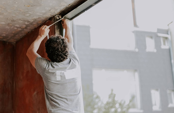 Ein junger kaukasischer Mann in grauem T-Shirt mit lockigem braunem Haar von hinten repariert eine Fensteröffnung, entfernt die Silikonisolierung und bereitet den Einbau eines Fensters mit Rahmen vor, Seitenansicht aus nächster Nähe. Das Konzept der Reparatur zu Hause, Wind - Foto, Bild