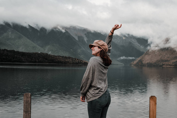 caucasian girl standing relaxed and happy smiling near lake wearing brown hat gray shirt and blue jeans with right hand stretched upwards with mountains in the background on a foggy day, nelson lakes - Photo, Image