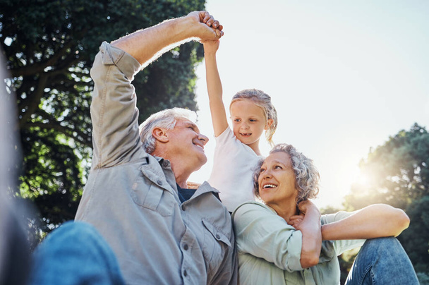 Grandparents playing together with a girl in the park in the morning. Family, love and grandchild bonding with grandmother and grandmother in a garden. Child holding hands with senior couple outside. - Фото, изображение