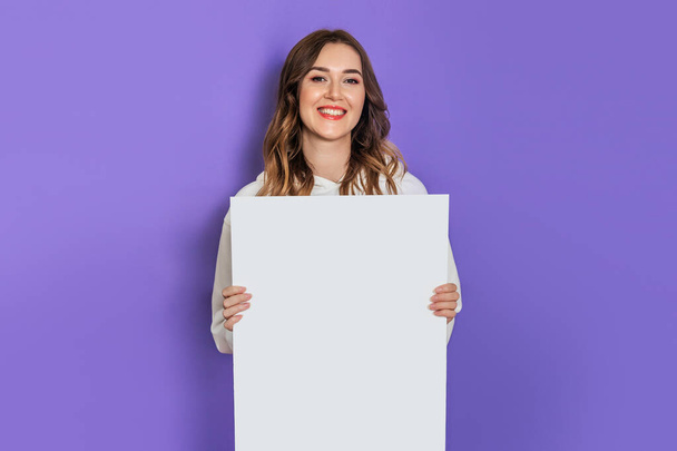 Νεαρή καυκάσια φοιτήτρια που κρατάει ένα λευκό φύλλο χαρτί, αφίσα, πλακάτ στα χέρια χαμογελώντας απομονωμένη στο λιλά φόντο. Αντιγραφή χώρου - Φωτογραφία, εικόνα