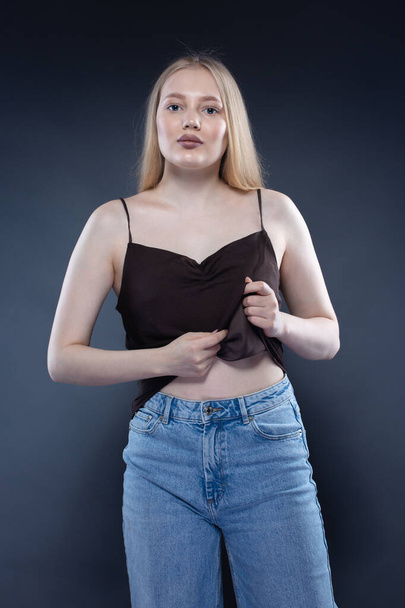 Young blonde woman in jeans and brown singlet on dark gray background - Фото, изображение