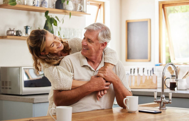 Senior couple, hug and bonding with morning coffee in a house or home kitchen in trust, security and love. Smile, happy and retirement elderly man and mature woman with tea and safety life insurance. - Photo, image