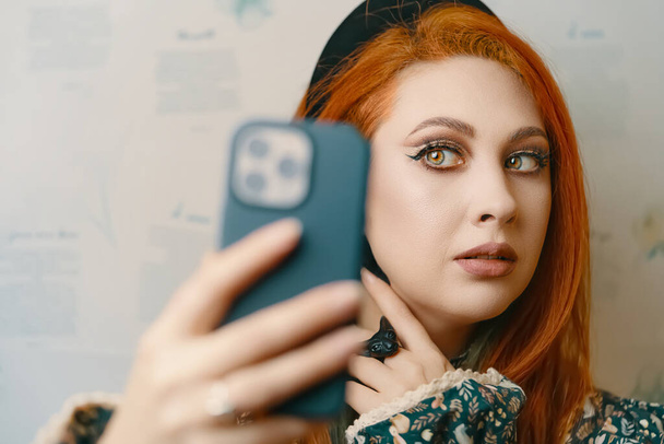 Mooi meisje neemt een selfie op de camera aan de voorkant van een moderne smartphone. Vrouwelijke blogger schiet zichzelf voor een blog over mode en make-up. Roodharige vrouw met een etnische slavische kroon op haar hoofd. - Foto, afbeelding