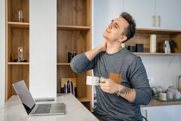 Un hombre en la cocina por la mañana sostenga el cuello con dolor de espalda dolorida que sufre de dolor de cuello mientras está de pie junto a la computadora portátil con taza de café exceso de trabajo y el concepto de agotamiento espacio de copia - Foto, imagen