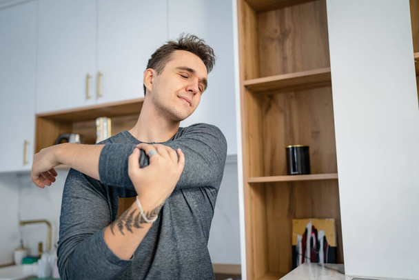 Een man in de keuken in de ochtend houden nek met pijn in de rug die lijden aan nekpijn terwijl staan door laptop computer met kopje koffie overwerk en uitputting concept kopieerruimte - Foto, afbeelding
