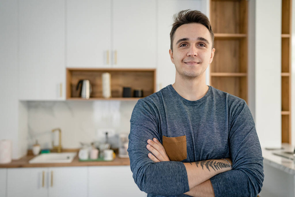 Vorderseite Porträt eines Mannes junge erwachsene kaukasische Mann steht in der Küche in Tag zu Hause glücklich lächeln zuversichtlich Kopie Raum Taille echte Menschen - Foto, Bild