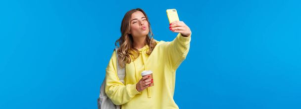 Cute positive and feminine beautiful young female student heading college holding paper cup from favorite cafe and backpack with books, taking selfie show kiss mwah face at mobile camera. - Foto, Imagen