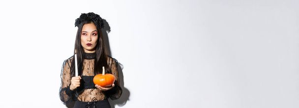 Image of asian woman in wicked witch costume, looking left serious, holding lit candle and pumpkin, celebrating halloween. - Photo, Image