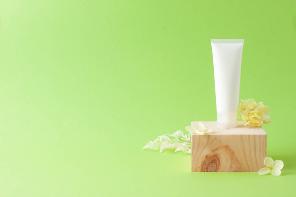 Tubo bianco di crema cosmetica con podio in legno e fiori su sfondo verde. Chiudi, copia spazio - Foto, immagini