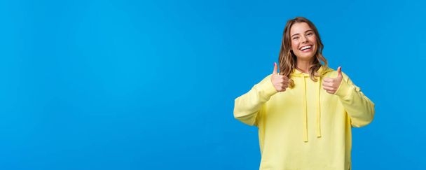 Sounds good Im in. Cheerful and optimistic good-looking young female blonde in yellow hoodie, show thumbs-up in approval, smiling nod acceptingly, like idea, blue background. - Photo, Image