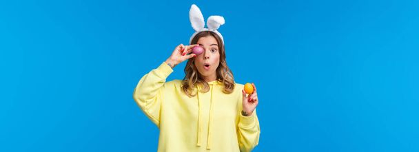Holidays, traditions and celebration concept. Amused lovely european female blonde in rabbit ears, look excited and surprised as holding two painted Easter day eggs, blue background. - Foto, Imagen
