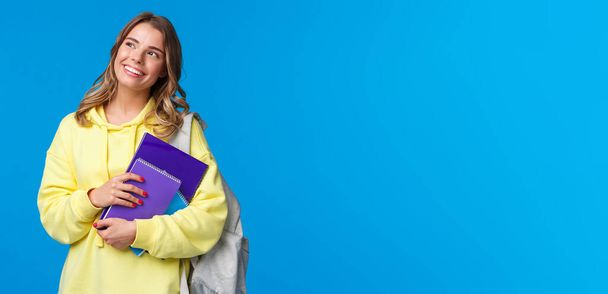 Ονειρεμένη χαρούμενη όμορφη ξανθιά κοπέλα που σπουδάζει στο κολέγιο, κρατάει σακίδιο και σημειωματάρια, το προσωπικό της εργασίας, κοιτάζει πάνω αριστερή γωνία στοχαστικό, στέκεται μπλε φόντο. - Φωτογραφία, εικόνα