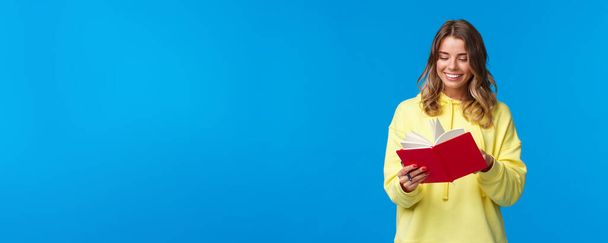 Girl bought new planner, writing down notes or lecture, smiling joyfully, holding red notebook, decide start secret diary, standing in yellow hoodie over blue background. - Foto, immagini
