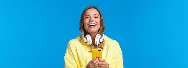 Retrato de cerca alegre chica rubia guapa con capucha amarilla y auriculares blancos, usando el teléfono móvil, escribiendo en su blog, compartiendo noticias con las redes sociales, mensajería y cámara de risa
. - Foto, imagen