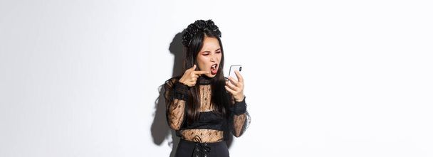 Portrét rozzlobené asijské ženy v čarodějnickém kostýmu hádající se na videohovoru, křičící na mobilní telefon a ukazující prstem, obviňující někoho, stojící nad bílým pozadím. - Fotografie, Obrázek