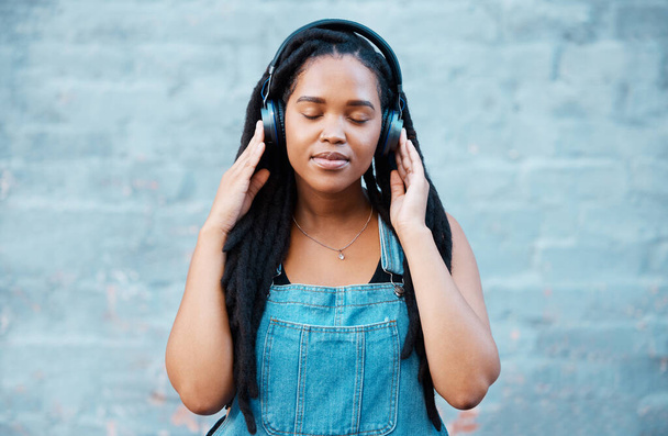 Czarna kobieta, słuchawki i muzyka z dziewczyną, która czuje się spokojna, zrelaksowana i zadowolona słuchając podcastu stojącego przed miastem lub murem miejskim. Afrykańska kobieta zrelaksowana podczas uważnego strumieniowania audio. - Zdjęcie, obraz