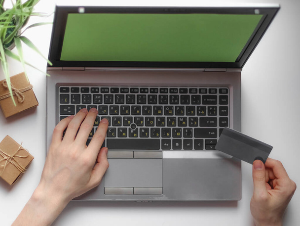 Χέρια ενός νεαρού καυκάσιου άνδρα που κρατά μια τραπεζική κάρτα στο χέρι του και πληκτρολογεί στο πληκτρολόγιο ενός φορητού υπολογιστή με μια πράσινη οθόνη, η οποία βρίσκεται σε μια λευκή επιφάνεια εργασίας με μικρά κουτιά δώρων χειροτεχνίας και ένα λουλούδι σε μια κατσαρόλα, κάθεται στο κρεβάτι στην κρεβατοκάμαρα, επίπεδη lay cl - Φωτογραφία, εικόνα