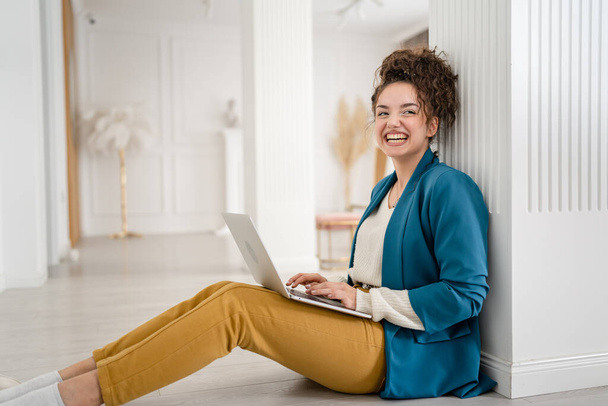 Genç bir kadın, beyaz kadın evde dizüstü bilgisayarla yerde oturuyor ve yan tarafa bakıyor mutlu bir gülüş, kıvırcık saçlı, yerli hayatı kopyalayan serbest bir esmer. - Fotoğraf, Görsel