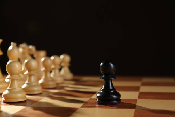 Black pawn in front of white pieces on wooden board against dark background. Competition concept - Foto, afbeelding