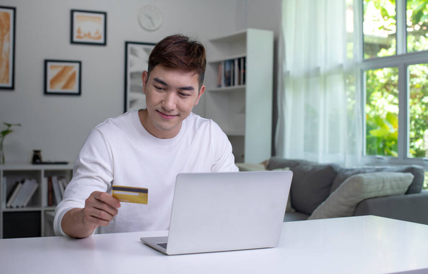 Mladý Asiat držící kreditní kartu a používající notebook. Podnikatel pracuje doma. Online nakupování, e-commerce, internetové bankovnictví, utrácení peněz, práce z domova - Fotografie, Obrázek