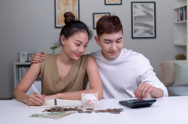 Retrato de una feliz pareja asiática en casa ahorrando dinero en una hucha. Mujer feliz y pareja de hombres en casa ahorrando dinero en una hucha y sonriendo. - Foto, Imagen