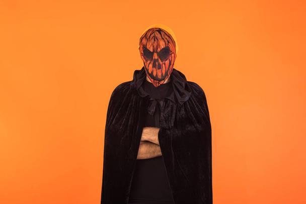Person with wearing Jack o Lantern pumpkin mask and black hooded cape, with arms crossed, celebrating Halloween, on orange background. Celebration concept, All Souls' Day and All Saints' Day. - Fotó, kép