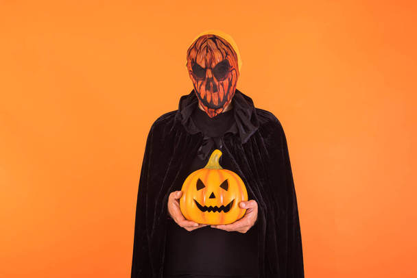 Πρόσωπο με μάσκα κολοκύθας Jack o Lantern και μαύρη κουκούλα κάπα, κρατώντας μια κολοκύθα, γιορτάζοντας το Halloween, σε πορτοκαλί φόντο. Εορταστική ιδέα, Ημέρα των Ψυχών και Ημέρα των Αγίων Πάντων. - Φωτογραφία, εικόνα